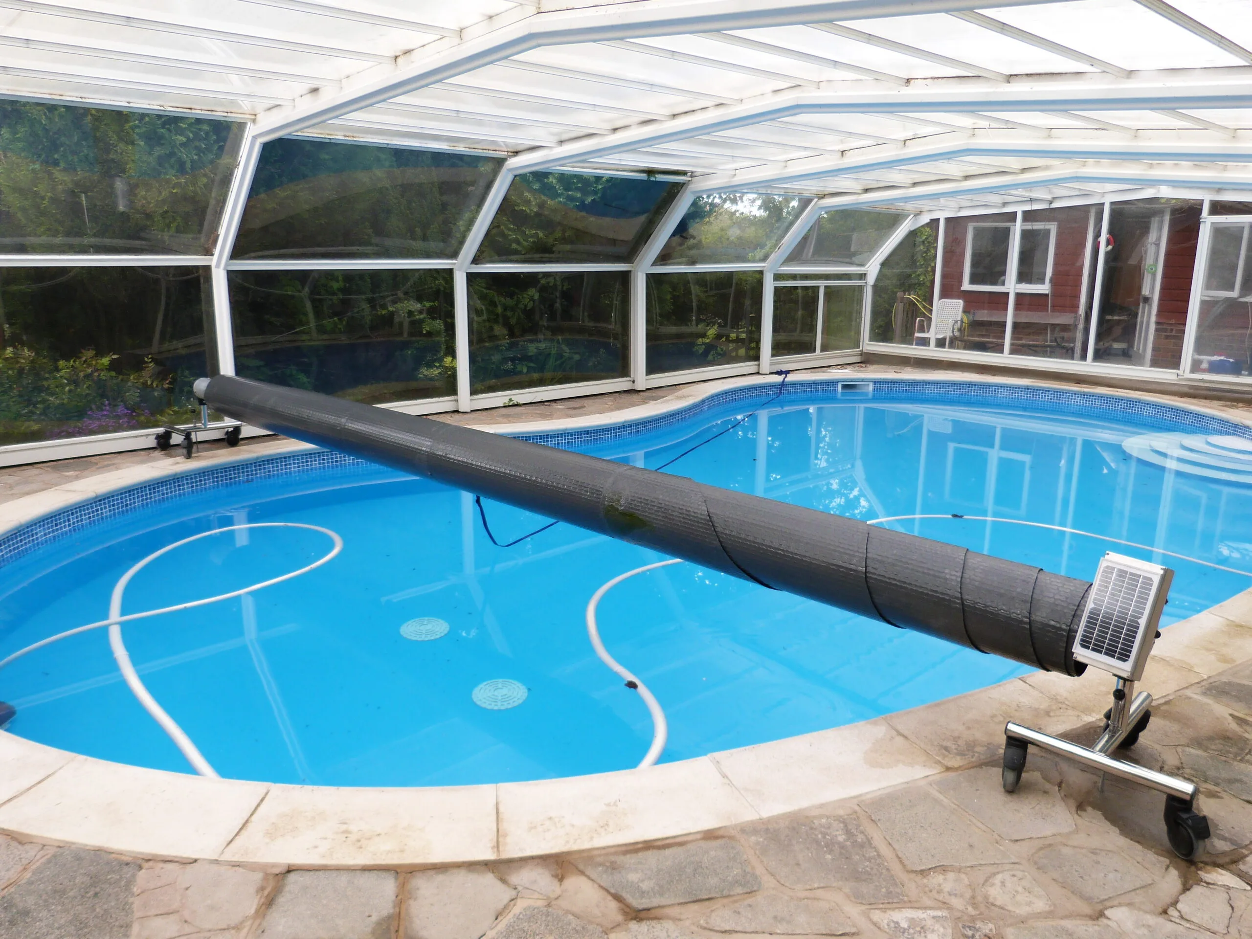Dark gray pool cover over a blue pool, enclosed by a clear glass structure. The pool is surrounded by stone. Solar panel.