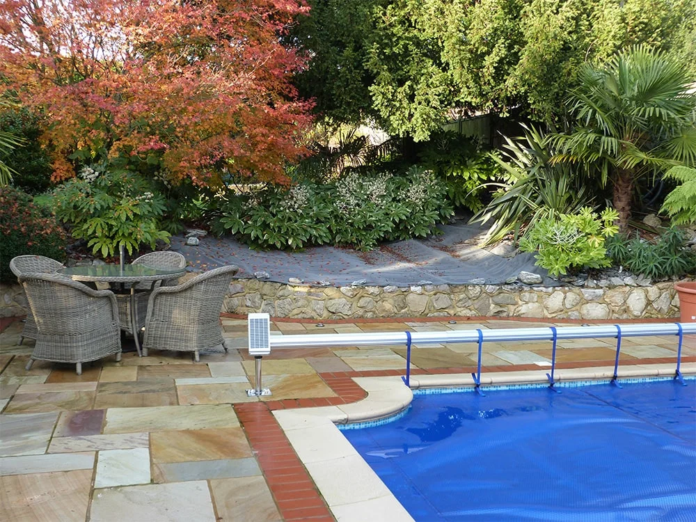 Blue solar pool cover on a reel with patio furniture. Grey wicker chairs and glass table on stone patio.